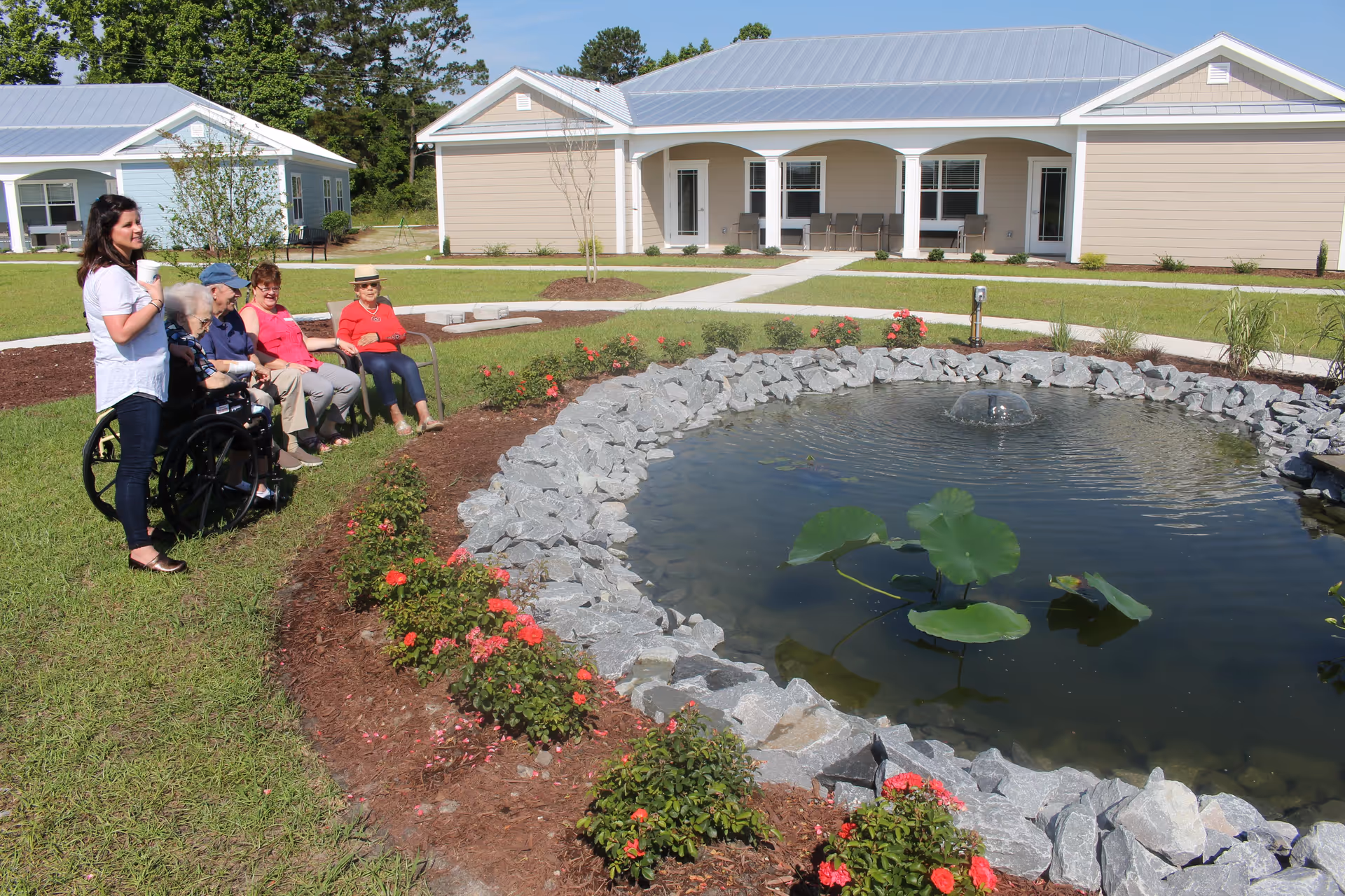 A group of elderly people sitting on benches and a woman standing beside them near a small pond with rocks and water lilies in a grassy outdoor area. Behind them are single-story buildings with light-colored siding and metal roofs under a clear blue sky.
