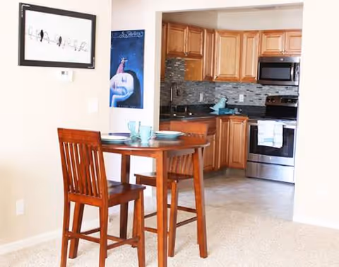 Wooden dining table with two chairs beside a kitchen with cabinets and stainless appliances