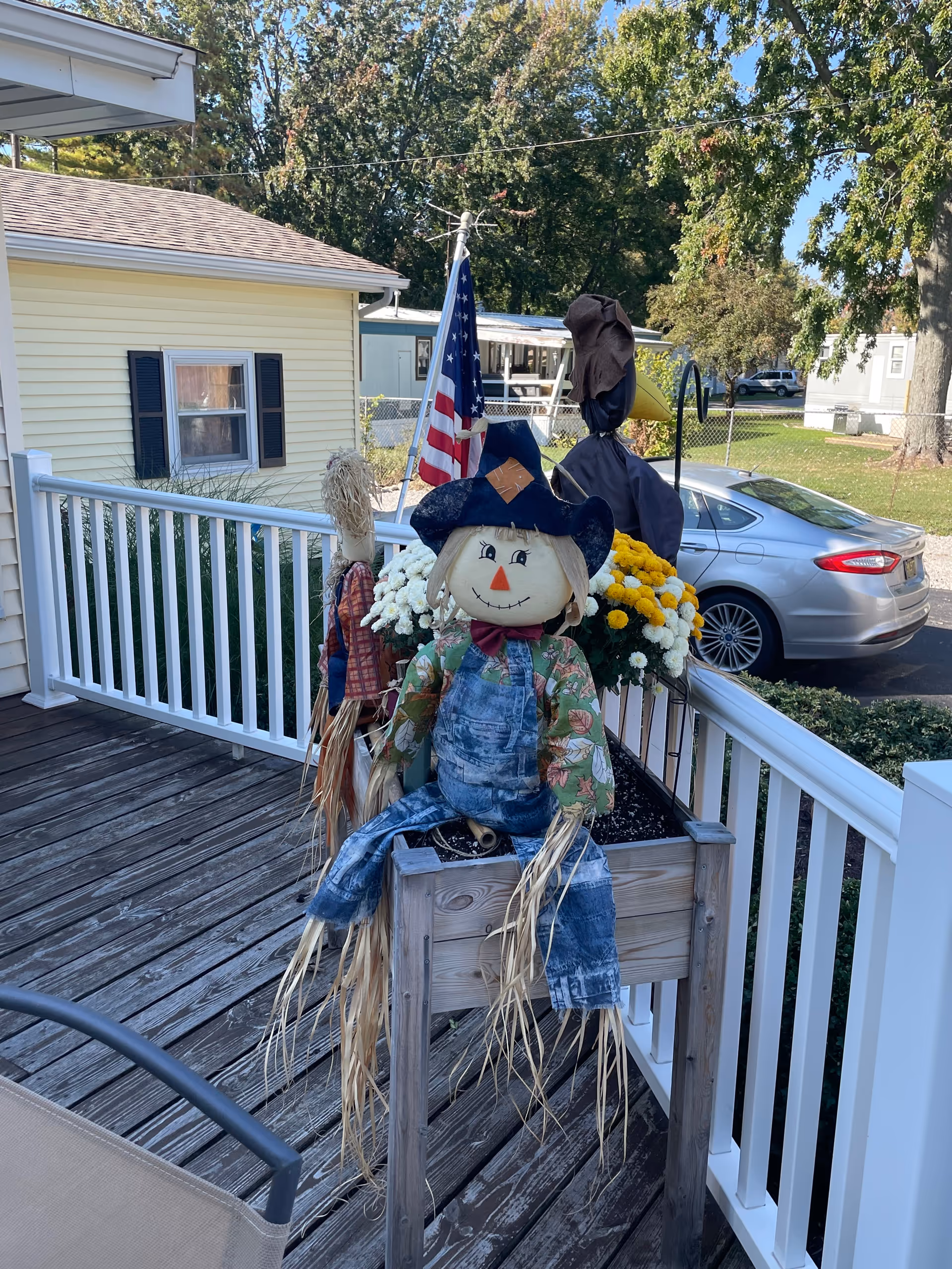 A wooden porch with white railing featuring a scarecrow decoration dressed in overalls and a hat sitting on a wooden planter box filled with yellow and white flowers. Behind the scarecrow is an American flag on a pole, a parked silver car, and a yellow house with black shutters. Trees and a chain-link fence are visible in the background.