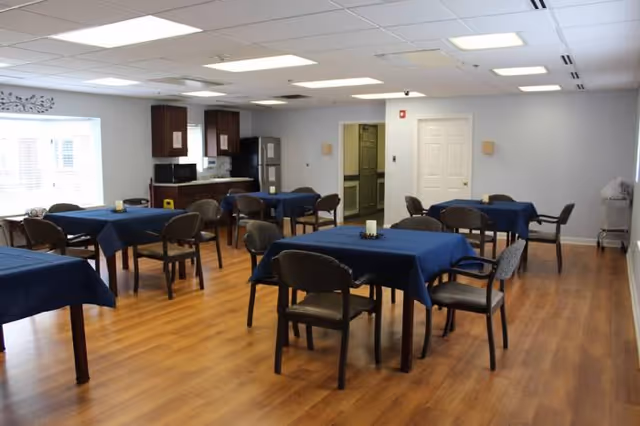 A dining room with several tables covered in dark blue tablecloths, each surrounded by four chairs. The room has wooden flooring, white walls, and a ceiling with recessed lighting. In the background, there is a small kitchen area with a refrigerator, microwave, and cabinets. A window with blinds is on the left side, and a door is visible on the far wall.