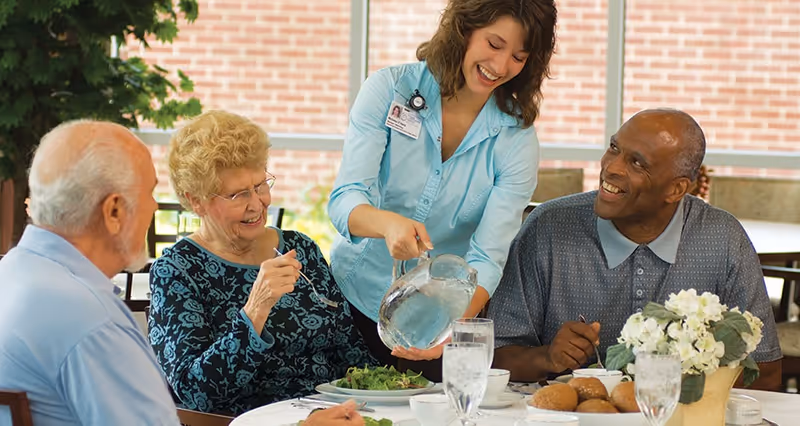 A caregiver pours water for three smiling elderly residents gathered around a dining table with plates, glasses, rolls, and a flower centerpiece.