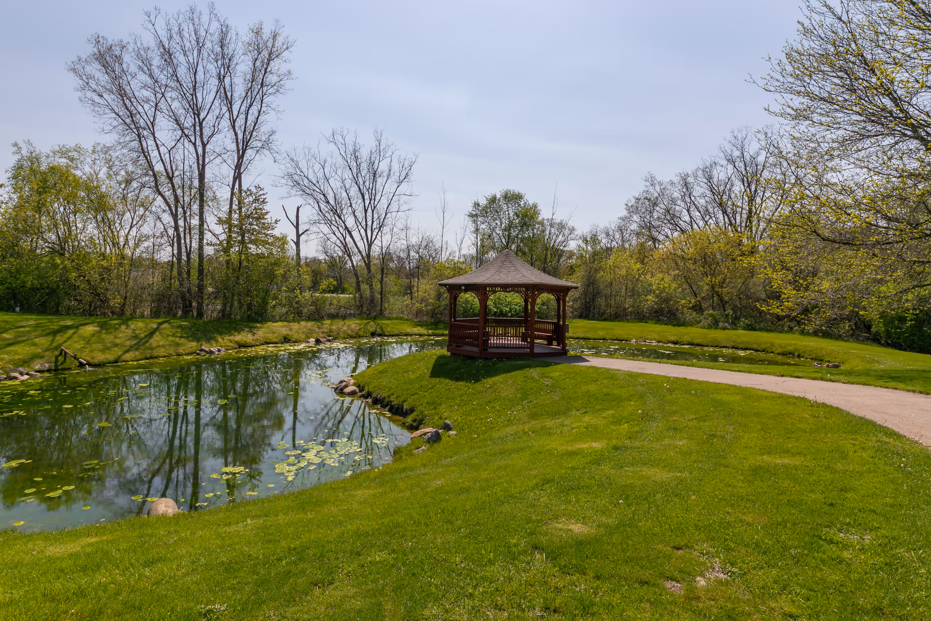 A peaceful outdoor scene featuring a small pond with lily pads, surrounded by green grass and trees. A wooden gazebo with a peaked roof is situated on a grassy area next to the pond, with a paved pathway leading to it. The sky is clear with light clouds.