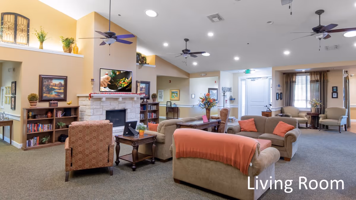 A spacious and well-lit living room area in an assisted living facility featuring multiple comfortable sofas and armchairs arranged around wooden coffee tables. The room has a stone fireplace with a TV mounted above it, ceiling fans, decorative plants, framed artwork on the walls, and large windows with curtains allowing natural light to enter. The carpeted floor and warm color tones create a cozy atmosphere.