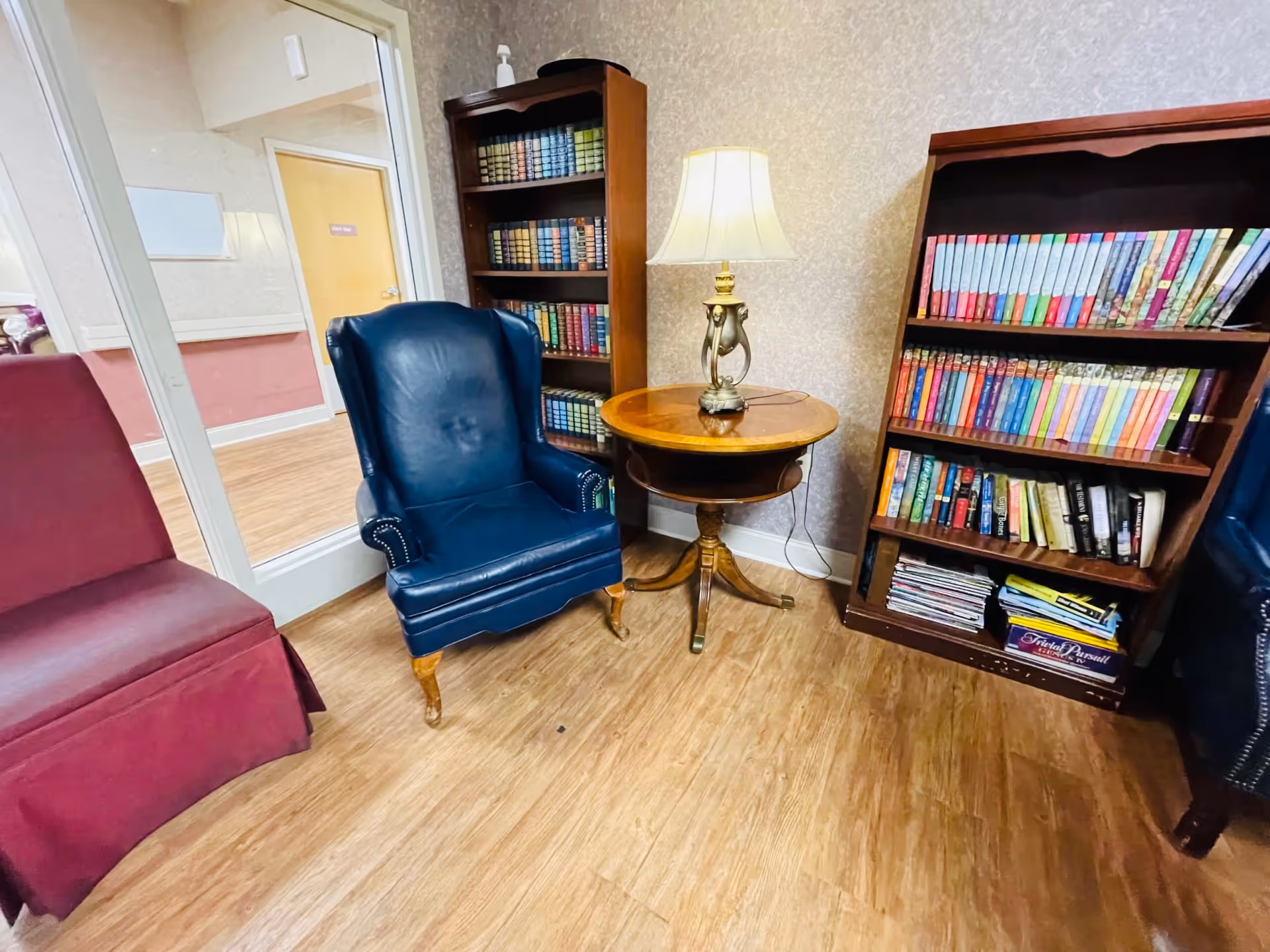 A cozy reading corner in a senior living facility featuring a blue leather armchair, a maroon upholstered chair, a round wooden side table with a decorative lamp, and two wooden bookshelves filled with books and magazines. The room has wood flooring and wallpapered walls.