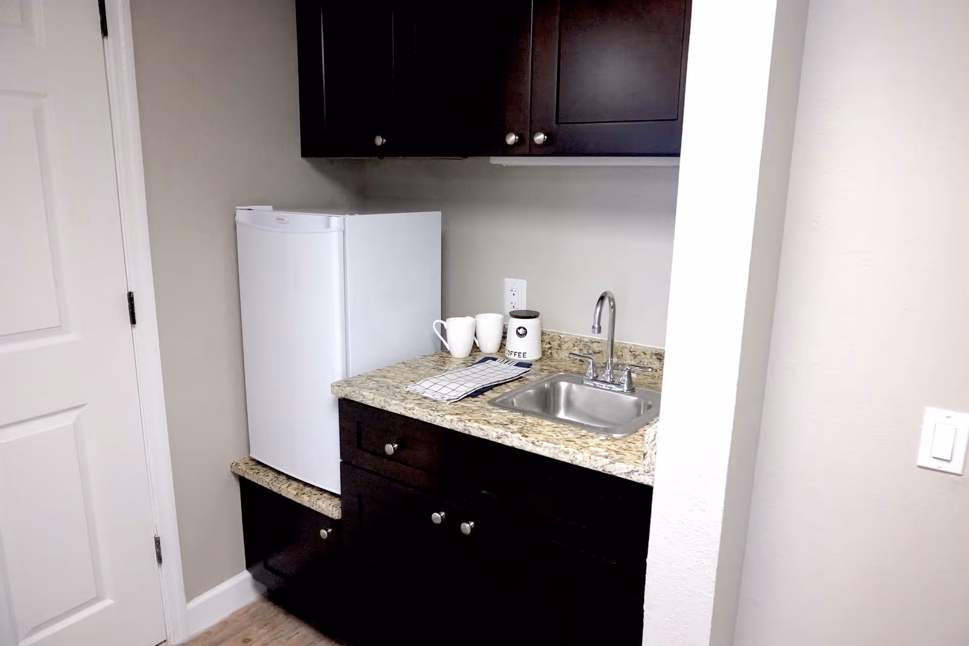 Small kitchenette area with dark wood cabinets, a granite countertop, a stainless steel sink with a faucet, a white mini refrigerator, two white mugs, a coffee container, and a folded checkered dish towel.