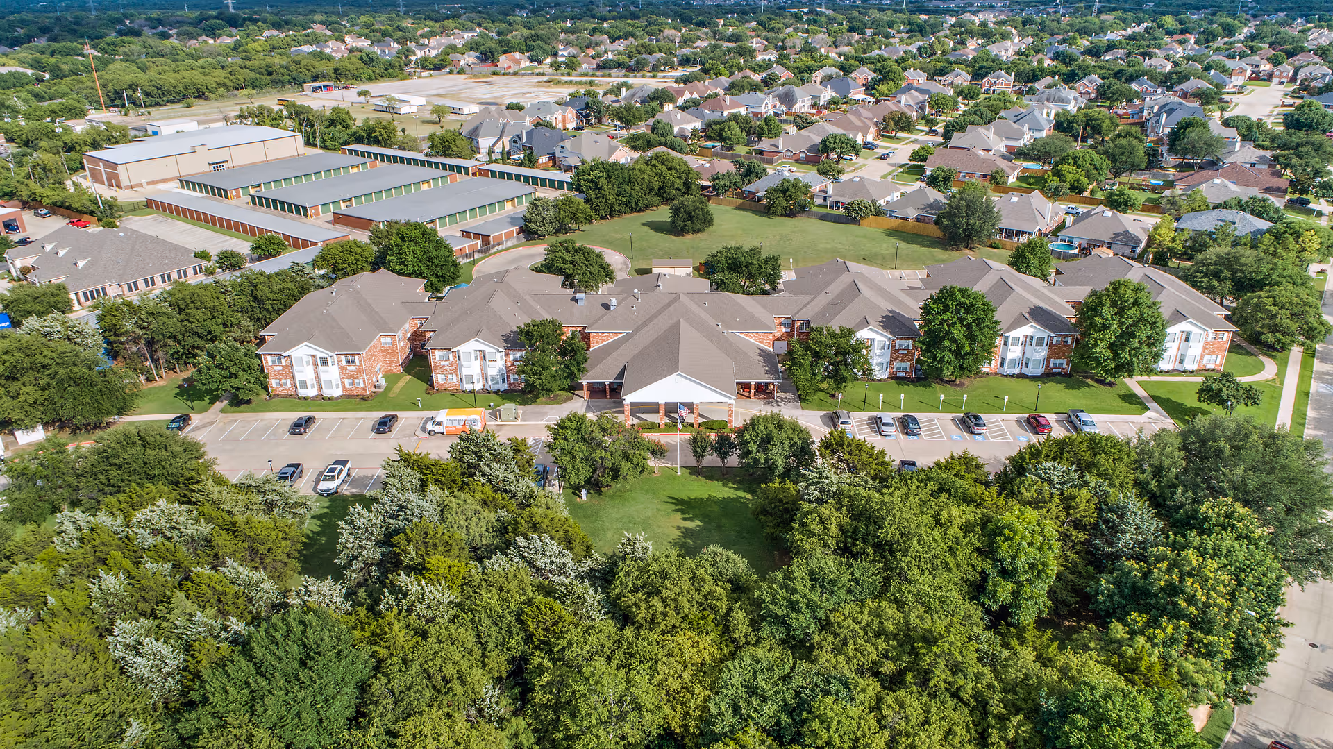 Aerial view of Morada Grand Prairie senior living facility showing multiple connected buildings with gray roofs and brick exteriors, surrounded by green trees and lawns. There is a parking lot with several cars in front of the buildings, and a residential neighborhood is visible in the background.