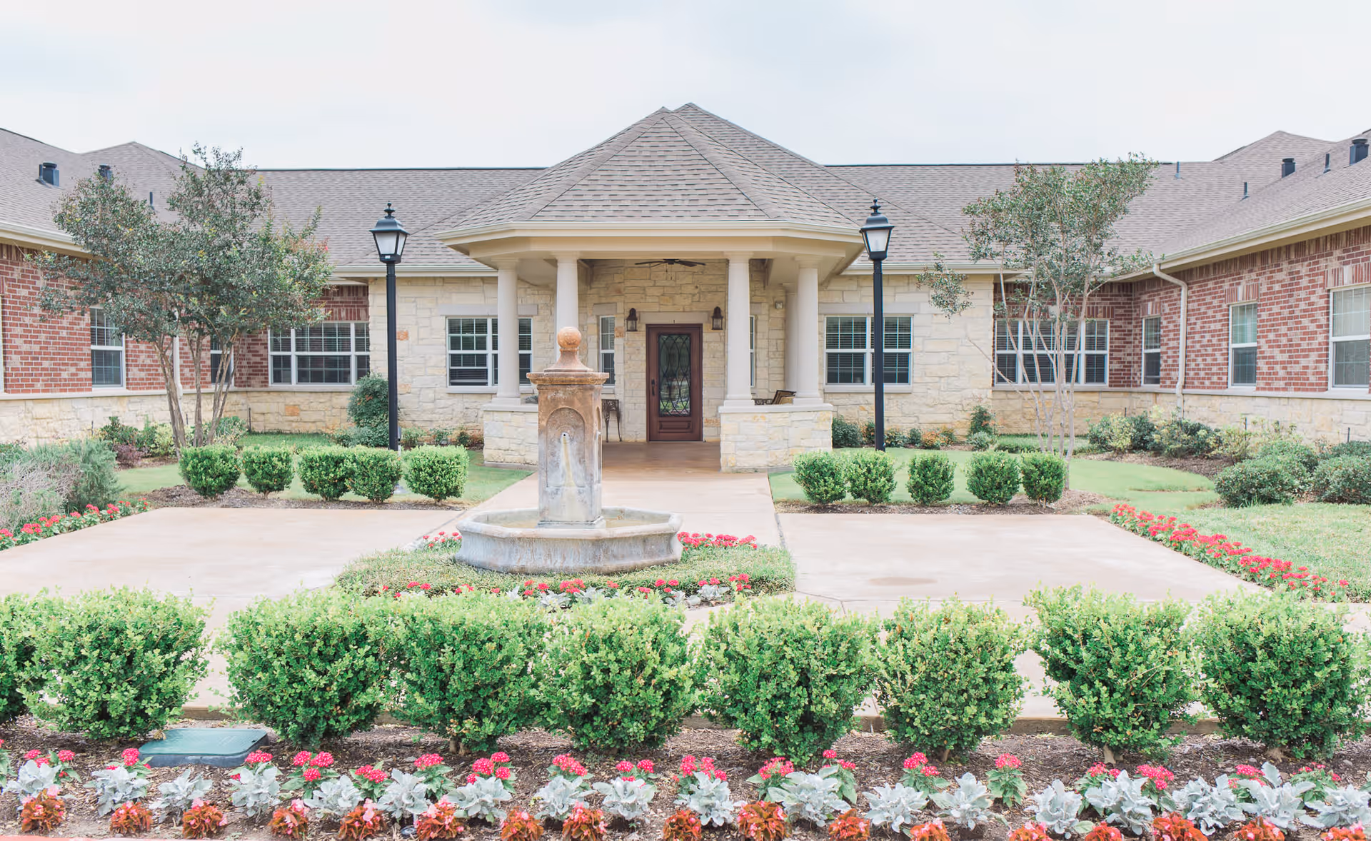 Front exterior view of The Heritage at Eldridge Parkway building with a covered entrance supported by columns, a central fountain, landscaped bushes, flowers, and two black lamp posts.