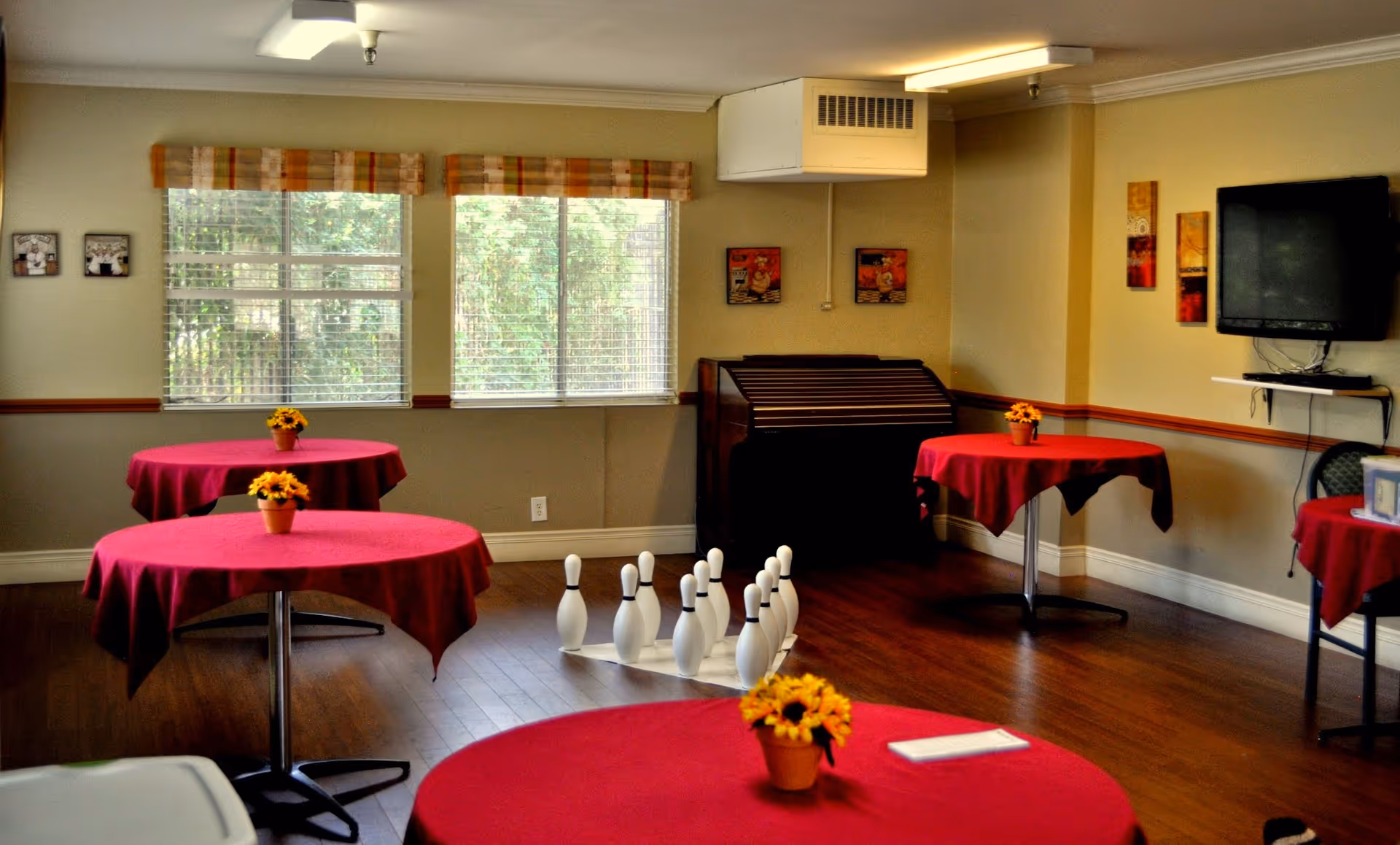 A common room with several round tables covered in red tablecloths, each decorated with a small pot of yellow flowers. There is a set of bowling pins arranged on the wooden floor in the center of the room. The room has large windows with blinds and plaid valances, a wall-mounted TV, and some framed pictures on the walls.