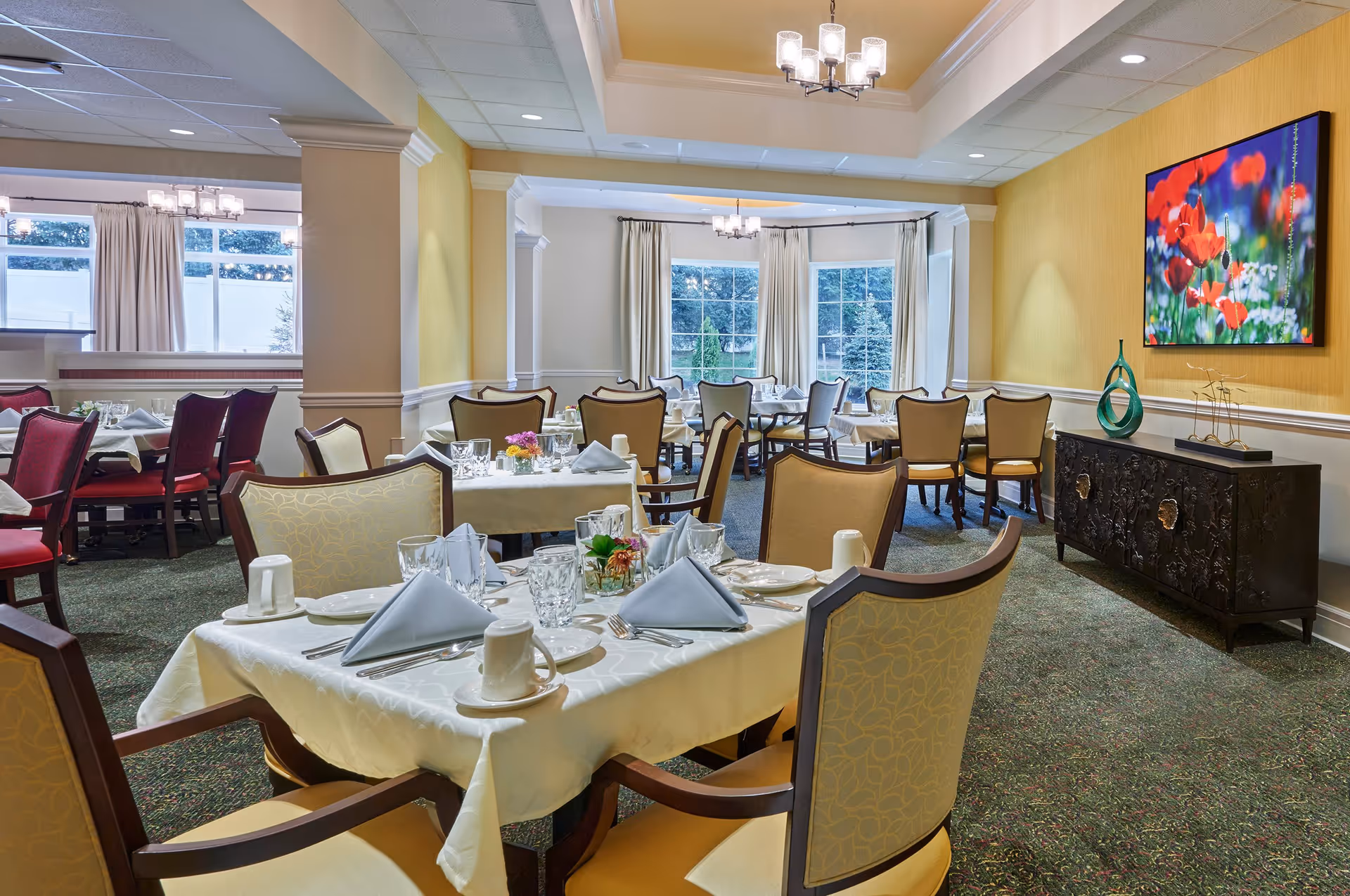 A well-lit dining room with multiple tables set with white tablecloths, folded napkins, glassware, and cups. The room features upholstered chairs in beige and red, large windows with cream curtains, a yellow accent wall with a colorful floral painting, and decorative vases on a dark wooden sideboard.