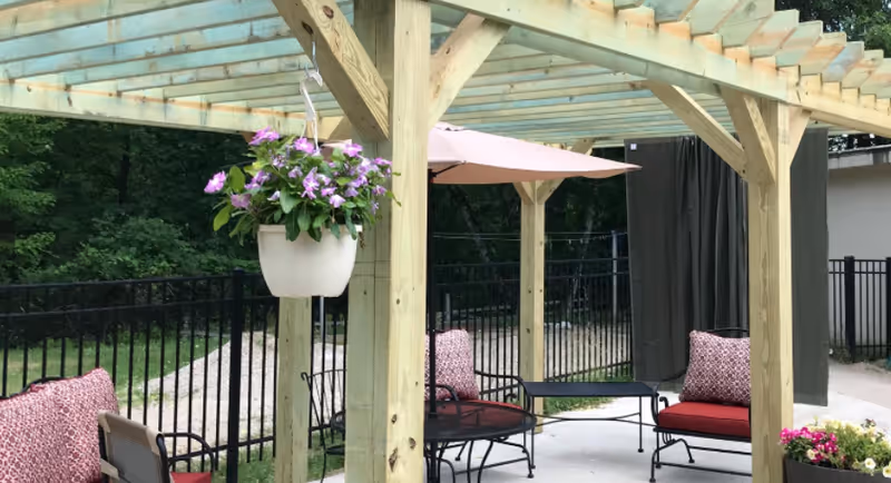 Outdoor seating area with a wooden pergola overhead, featuring cushioned chairs and a table. There is a hanging white planter with purple flowers attached to one of the pergola posts. A pink umbrella provides shade over part of the seating area. The space is enclosed by a black metal fence and surrounded by greenery and trees.