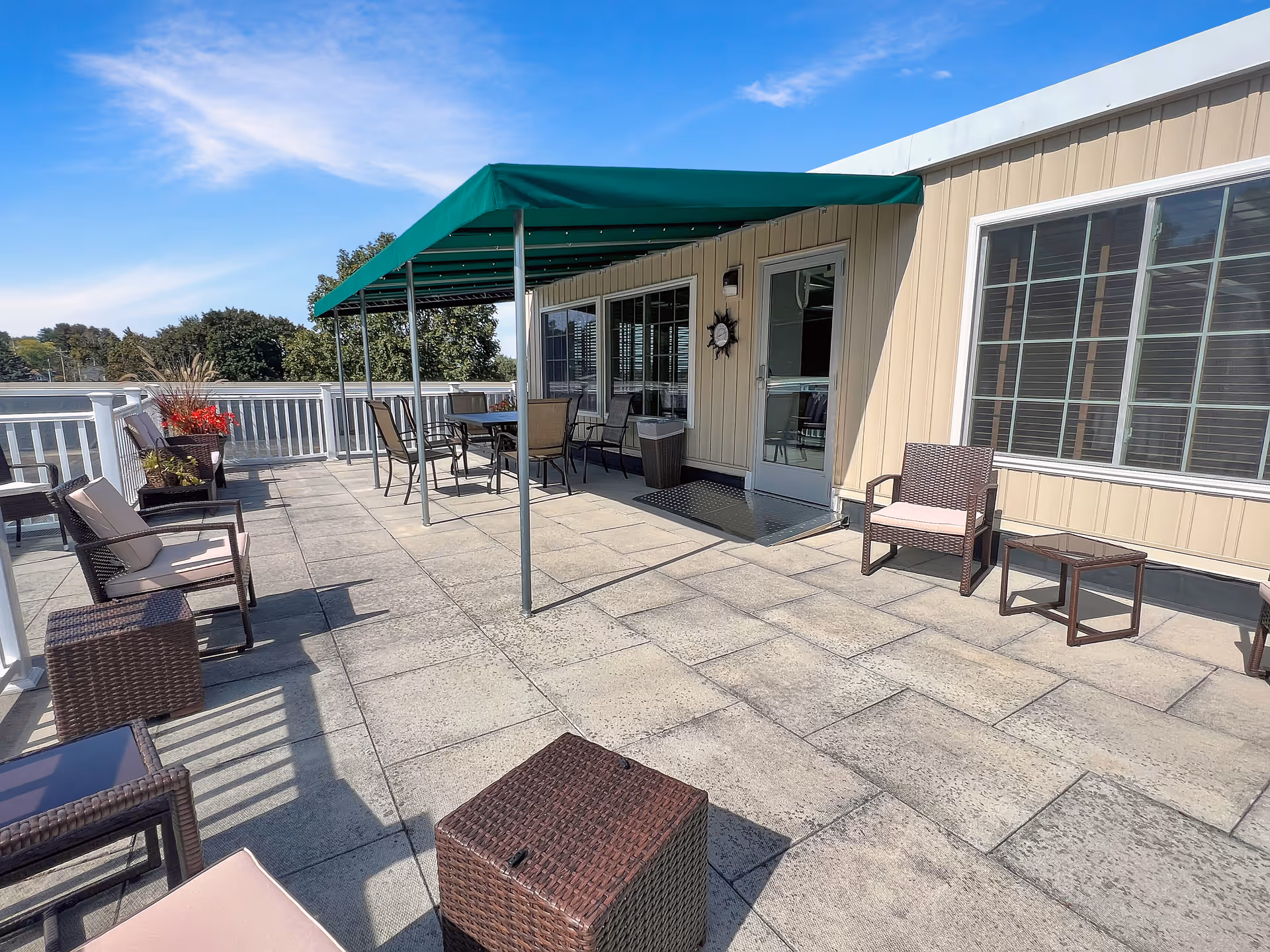 Outdoor patio area with several cushioned wicker chairs and small tables arranged on a tiled floor. A green canopy provides shade over a dining table with chairs near a building with beige siding and large windows. There are plants in pots along the white railing, and trees are visible in the background under a clear blue sky.