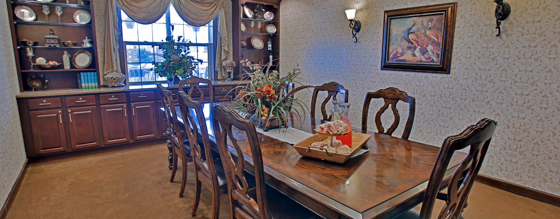 A formal dining room with a long wooden table surrounded by eight matching wooden chairs. The table is decorated with a floral centerpiece and a wooden tray holding a glass pitcher with red liquid and some pastries. The room has patterned wallpaper, a large window with draped curtains, built-in wooden cabinets with decorative plates and glassware, and a framed painting of peacocks on the wall.