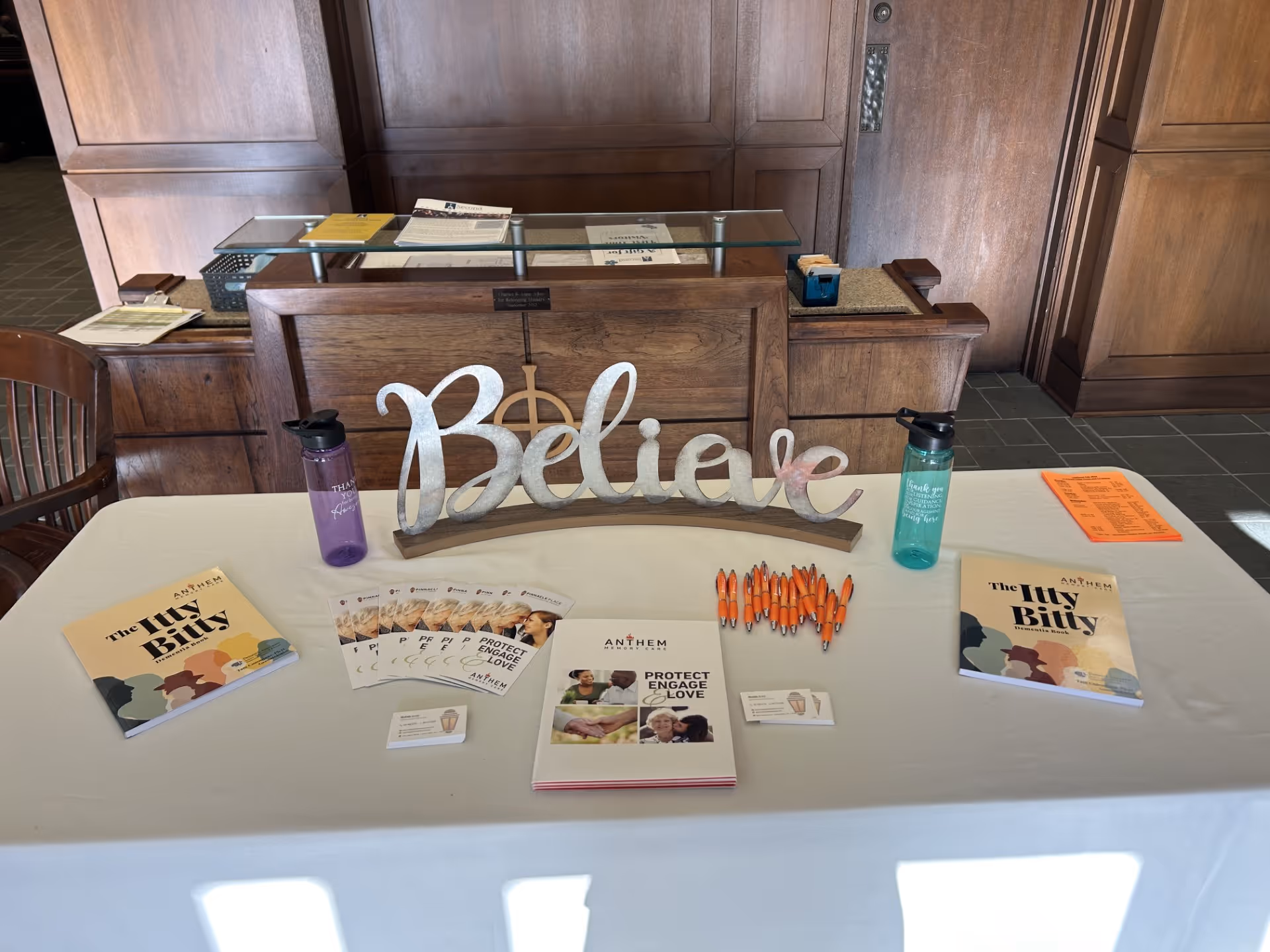 A table covered with a white tablecloth displaying brochures, booklets titled 'The Itsy Bitsy', orange pens, business cards, and two water bottles. A decorative wooden sign spelling 'Believe' is placed in the center of the table. Behind the table is a wooden counter and a door.