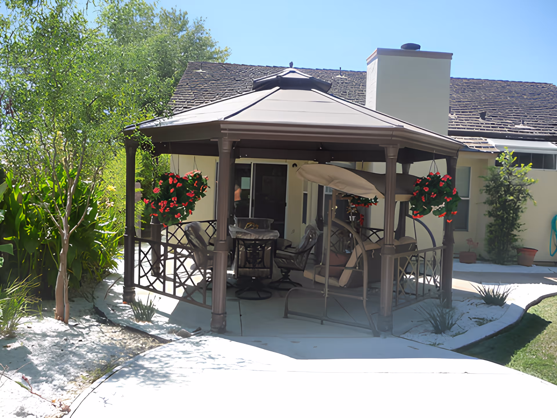 A covered outdoor gazebo on a patio with hanging flower baskets, seating and a swing in front of a house.