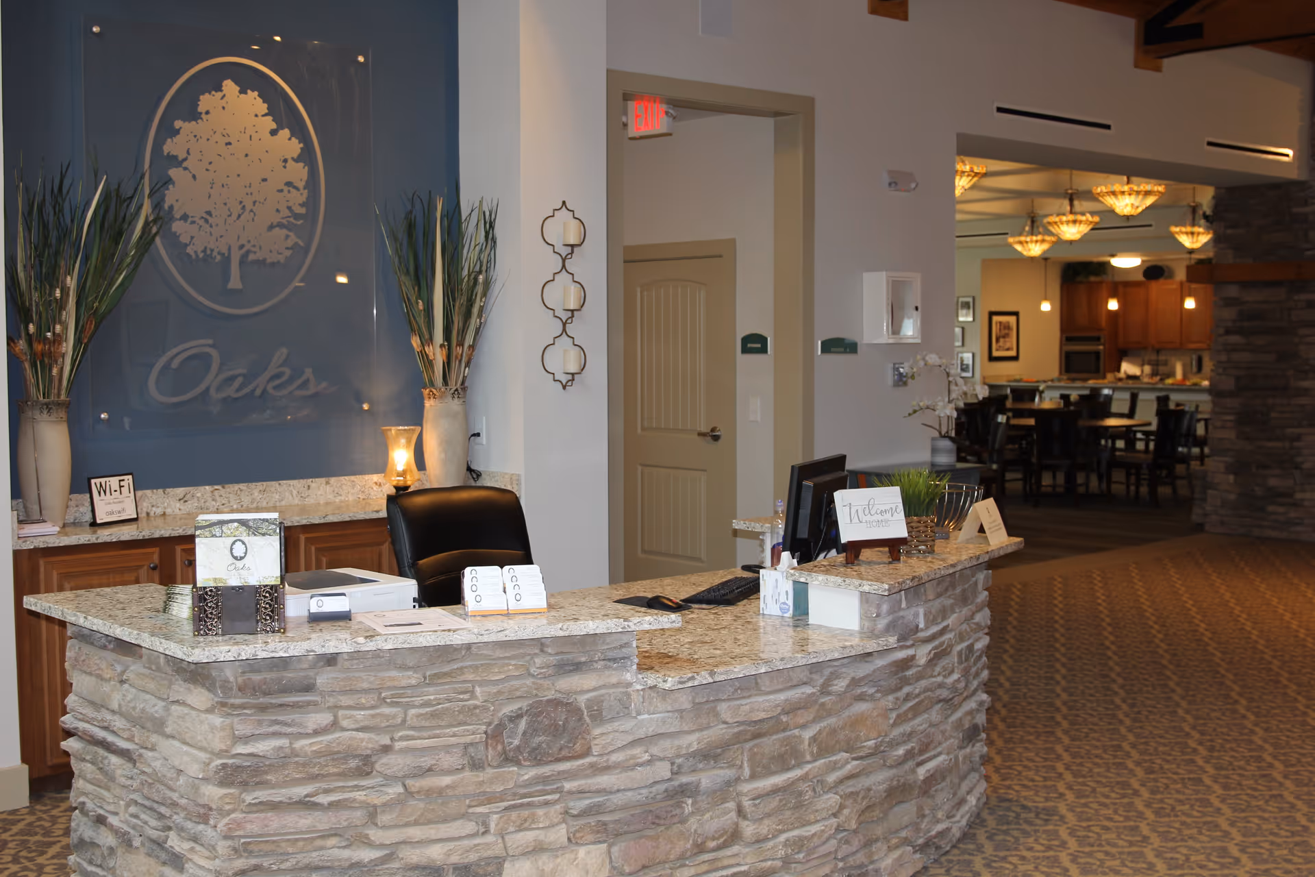 Reception desk area inside Oaks at Grove Park facility with a stone facade counter, a black office chair, computer, and decorative plants. Behind the desk is a large sign with a tree logo and the word 'Oaks'. In the background, there is a dining area with tables, chairs, and hanging light fixtures.