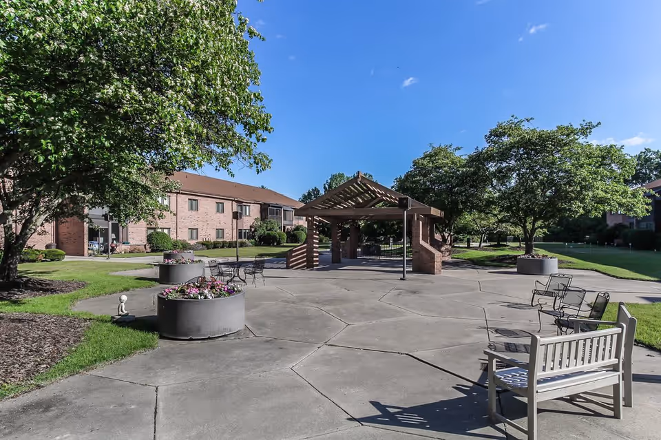 Outdoor courtyard area at Porter Hills Village featuring a paved open space with metal chairs and benches, large planters with flowers, a wooden pergola structure, and surrounding green lawns with trees under a clear blue sky.