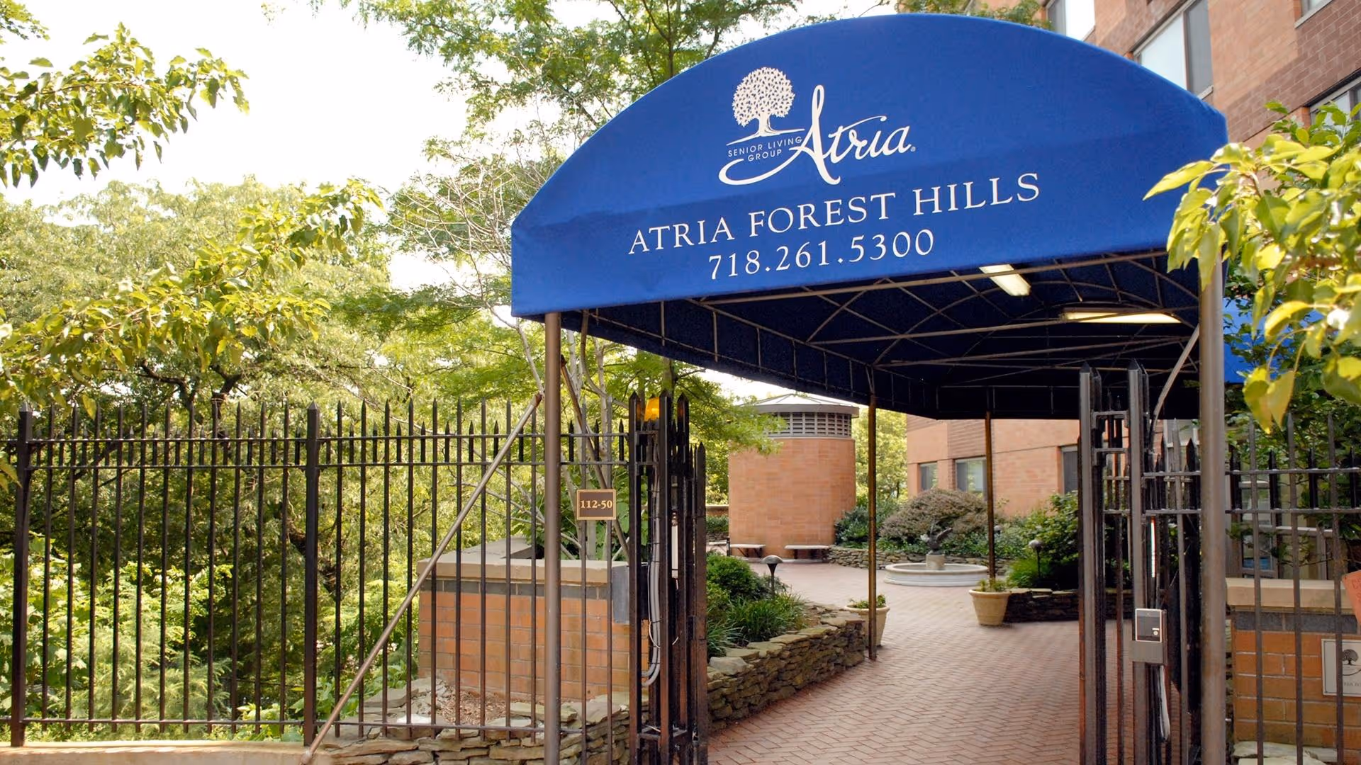 Entrance to Atria Forest Hills senior living facility with a blue canopy displaying the facility name and phone number, surrounded by greenery and a brick pathway leading inside.