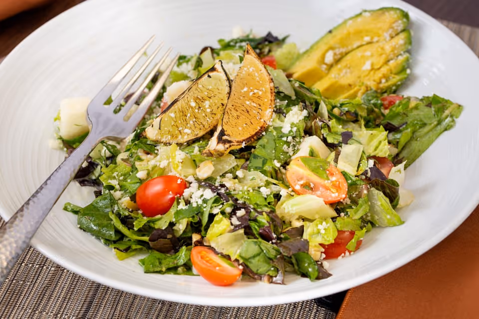 Close-up of a fresh salad with cherry tomatoes, avocado slices and grilled lemon wedges in a white bowl with a fork.