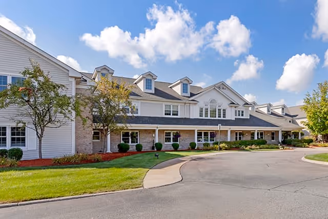 Exterior view of a two-story senior living facility building with white siding and stone accents, surrounded by green lawns, small trees, and a curved driveway under a blue sky with scattered clouds.