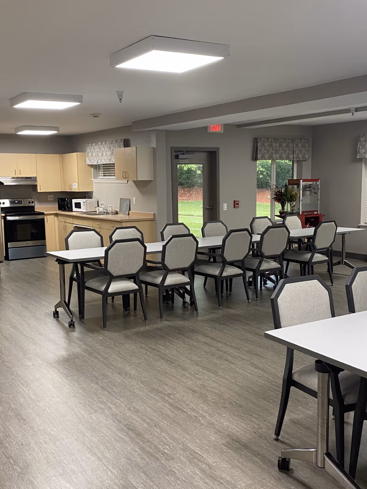 A clean and modern dining area with several tables and chairs arranged in rows. The room has light wood flooring and neutral-colored walls. In the background, there is a kitchen area with light wood cabinets, a stove, microwave, and sink. Large windows and a door provide natural light and a view of greenery outside. A popcorn machine and a vase with flowers are visible near the windows.