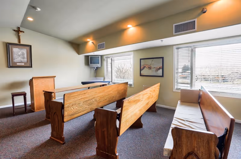 A small chapel-like interior with wooden pews, a lectern, a wall crucifix and windows letting in natural light.