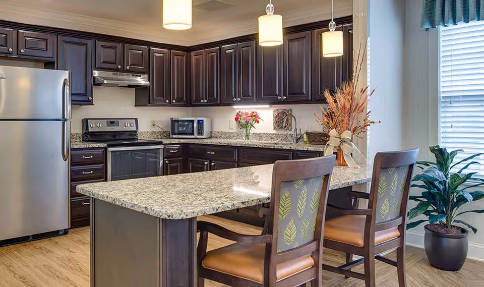 Kitchen with a granite island, dark wood cabinets, stainless steel appliances, pendant lights, and two chairs.