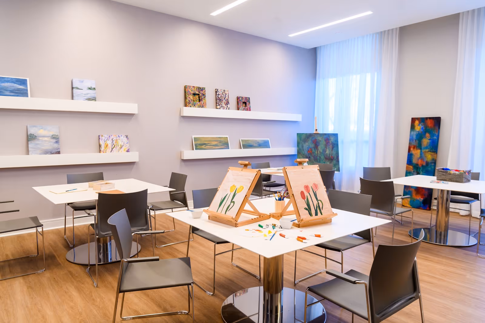 A bright art activity room with several tables and chairs arranged neatly. Two tables in the foreground have easels with paintings of tulips, paintbrushes, and art supplies. The walls display various colorful paintings on floating shelves and an easel near a large window with sheer white curtains. The floor is wooden, and the room has a clean, modern design.