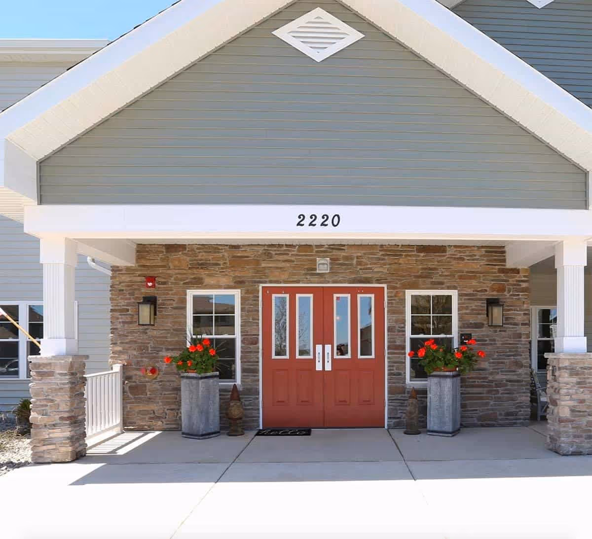 Covered building entrance with red double doors, stone facade, the number 2220 above the porch, and potted red flowers.