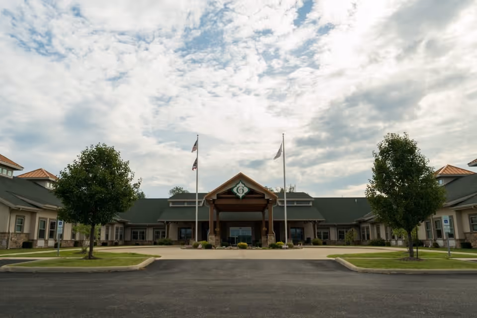 Front exterior view of The Gables of Canton facility with a driveway leading to the entrance, two flagpoles with flags, and trees on either side of the driveway under a partly cloudy sky.