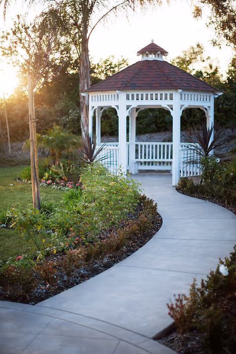 A white wooden gazebo with a red shingled roof situated at the end of a curved concrete pathway surrounded by green plants, flowers, and trees in a garden setting during sunset.