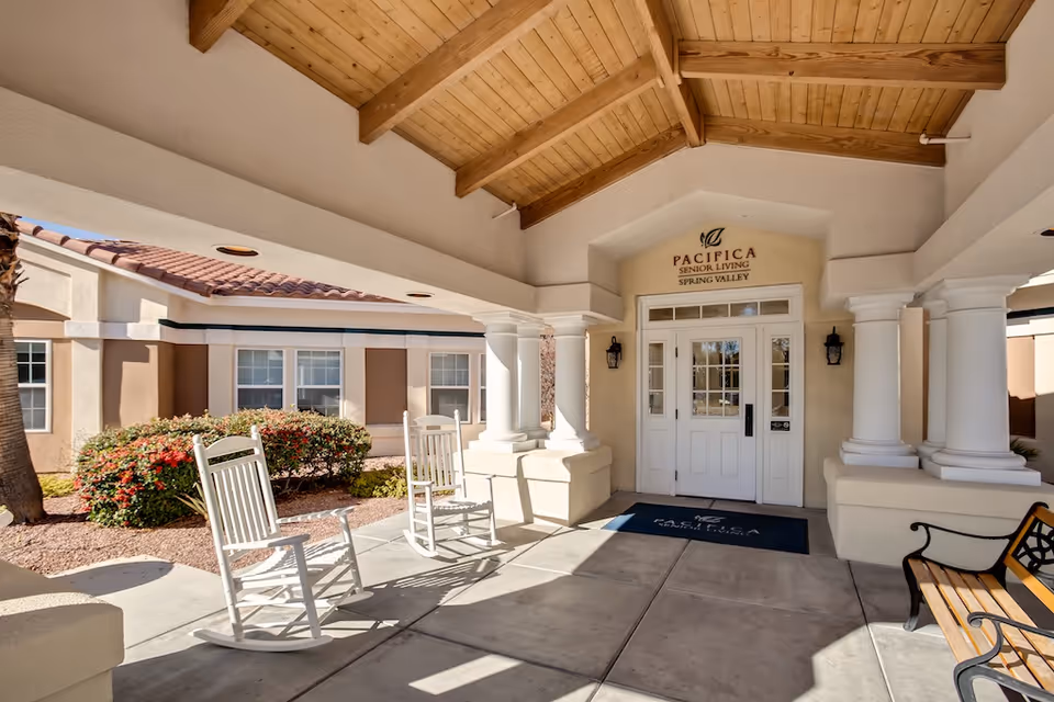 Covered entrance to Pacifica Senior Living Spring Valley with white rocking chairs, columns, and a bench.