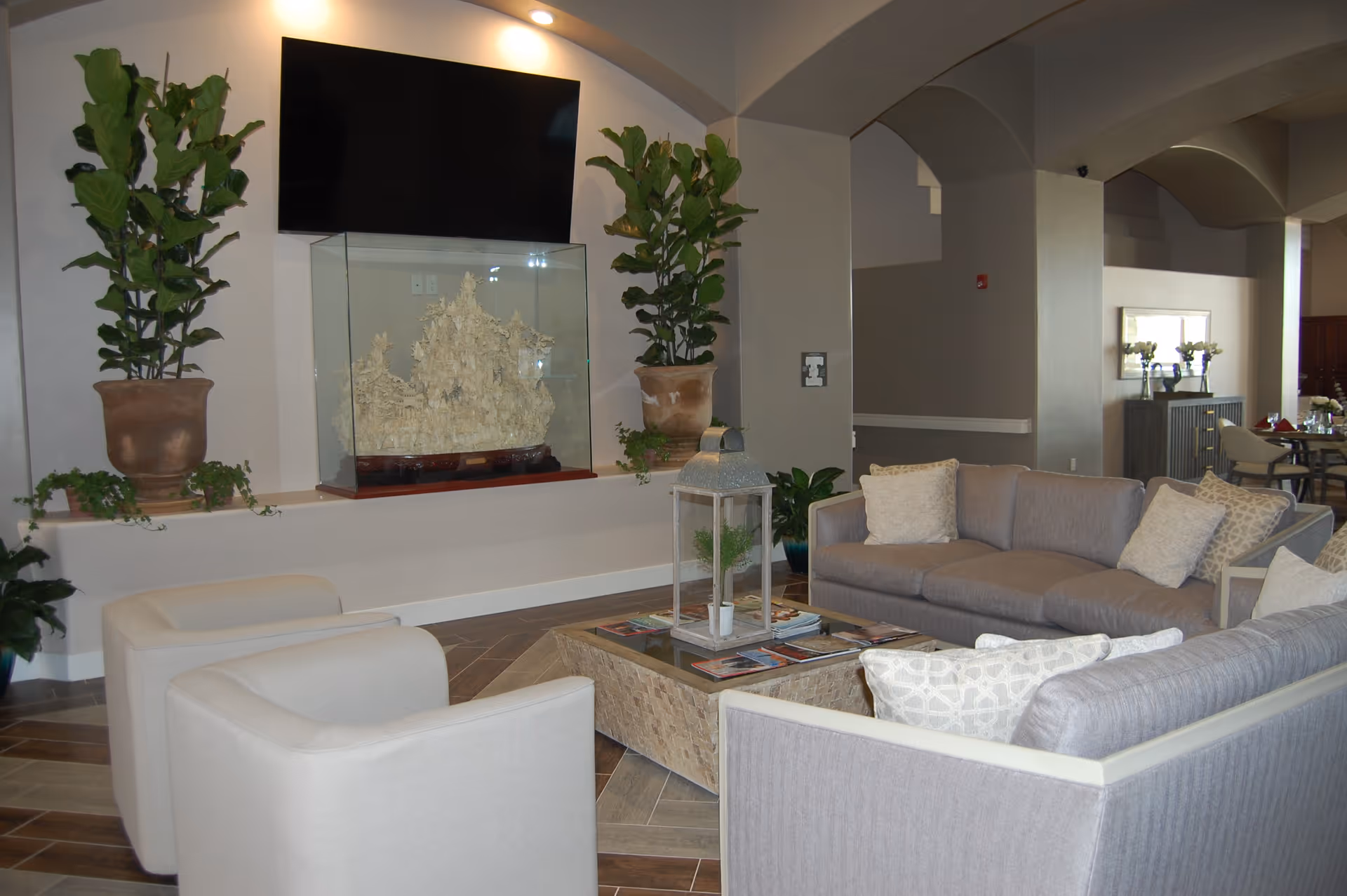 A modern living room area with two beige armchairs and two gray sofas arranged around a rectangular coffee table. The coffee table has a glass top and a decorative lantern with a small plant inside. Behind the seating area, there are two large potted plants flanking a glass display case with a carved sculpture inside. Above the display case is a large flat-screen TV mounted on the wall. The room has neutral tones with tiled flooring and soft lighting.