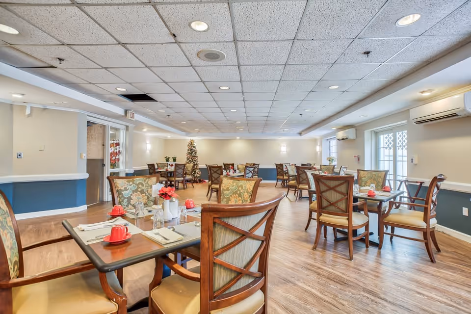 A spacious dining room with multiple wooden tables and chairs arranged neatly. Each table is set with red cups, glasses, napkins, and small flower vases. The room has a light-colored ceiling with recessed lighting, wood flooring, and light-colored walls with blue accents. A decorated Christmas tree is visible in the background near the far wall.