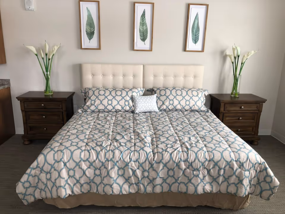Neatly made bed with patterned bedding, a tufted headboard, matching nightstands with vases of calla lilies, and three framed leaf prints above.