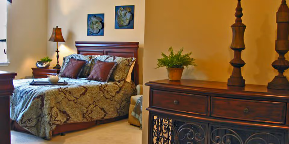 A cozy bedroom with a wooden headboard bed covered in a patterned bedspread and two brown pillows. There is a bedside table with a lamp and a small plant, two framed floral artworks on the wall above the bed, and a wooden dresser with decorative items and a potted plant.