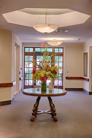 A hallway in a senior living facility with a round wooden table in the center holding a large floral arrangement. The hallway has beige walls, carpeted floor, and two ceiling light fixtures. At the end of the hallway, there is a set of glass doors with wooden frames letting in natural light.