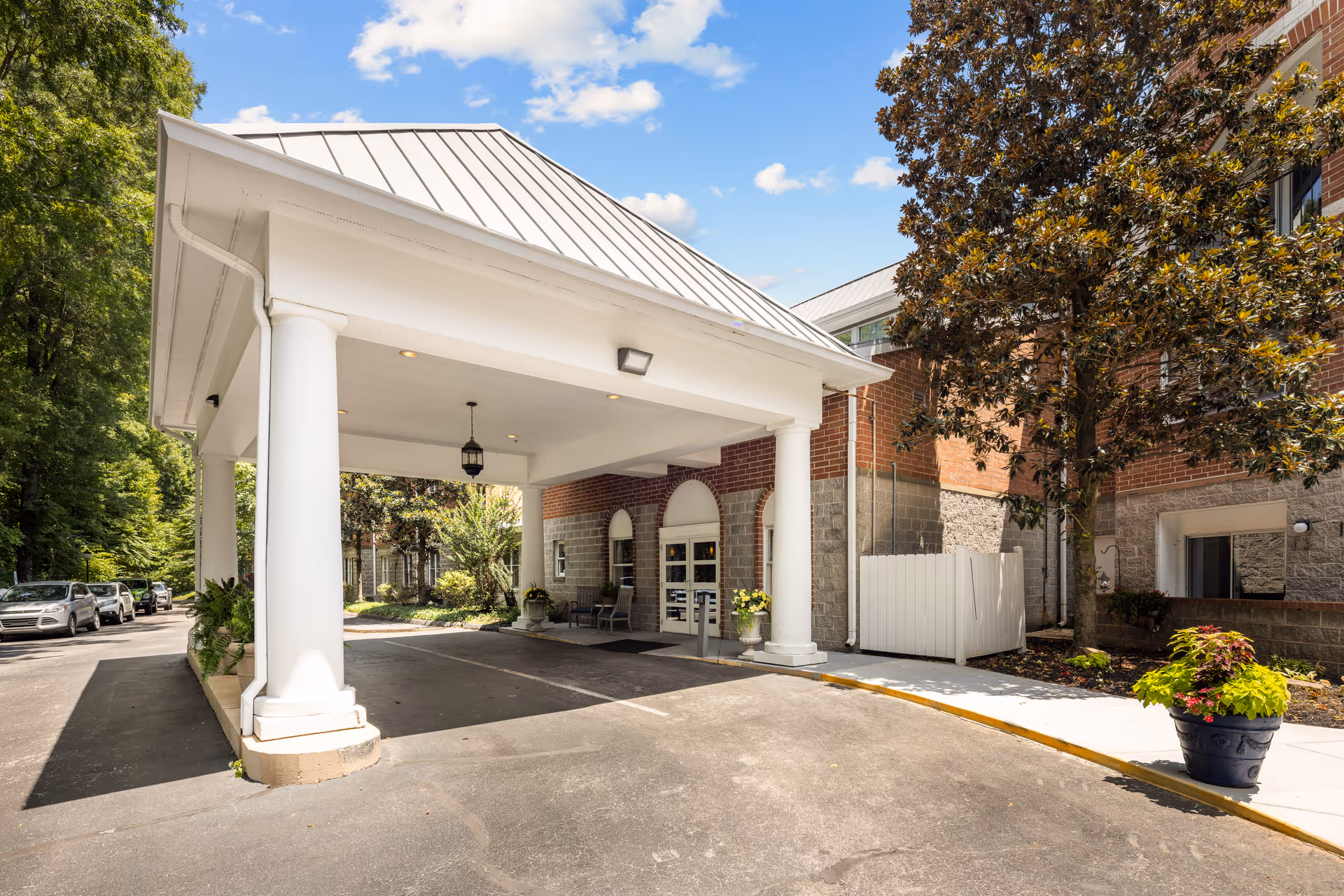 Entrance to a senior living facility with a covered drop-off area supported by white columns. The building has brick and stone exterior walls, with a tree and potted plants near the entrance. Several parked cars are visible along the driveway on the left side.