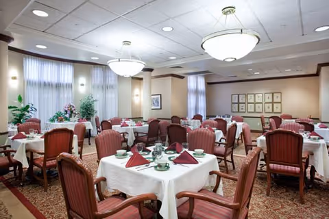 Bright communal dining room with round tables set with white tablecloths, folded burgundy napkins, cups and saucers, and upholstered chairs under chandeliers.