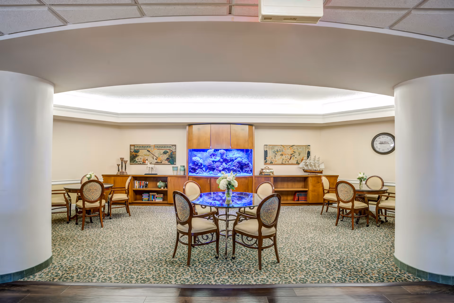 A well-lit common area with round tables and upholstered chairs arranged on a patterned carpet. In the background, there is a large built-in aquarium with blue lighting, flanked by wooden cabinets and decorative items including a model ship, candlesticks, and framed artwork. Two large white columns frame the space.