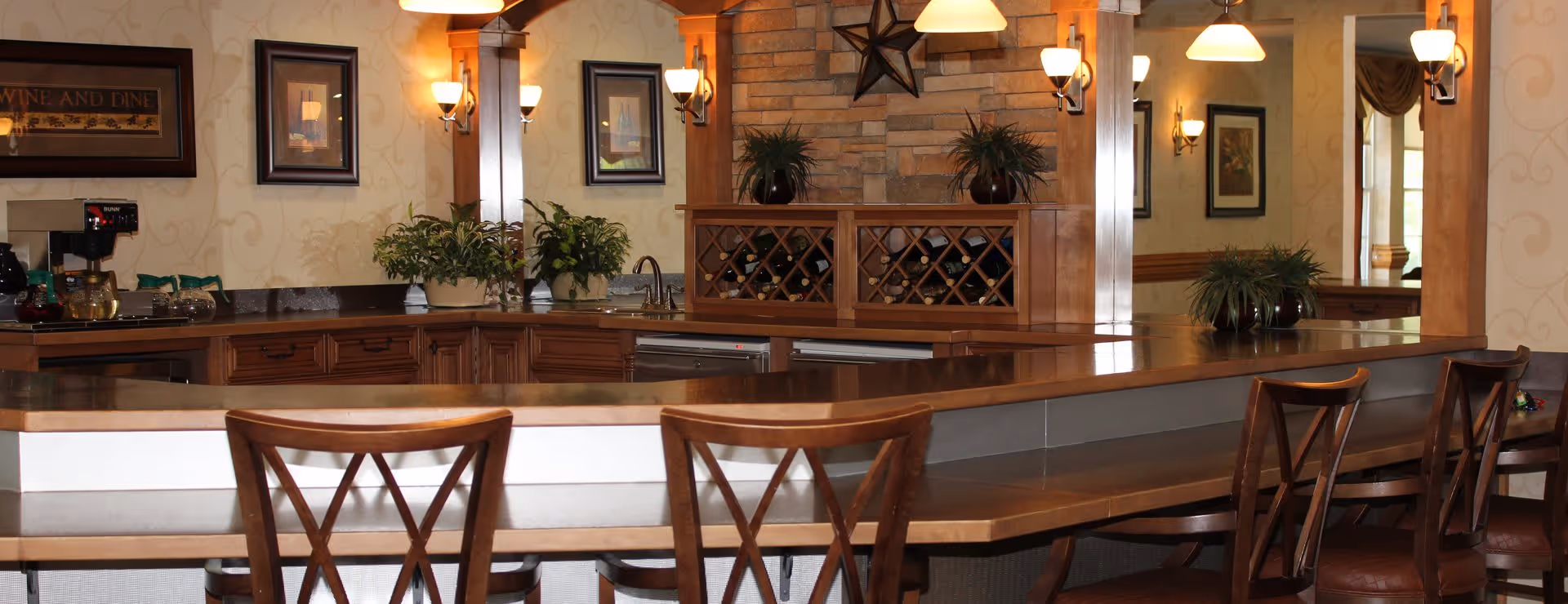Interior view of a warm, inviting dining or bar area with wooden chairs lined up along a counter. The background features a wine rack, plants, framed pictures on the wall, and soft lighting from wall sconces and hanging lamps.