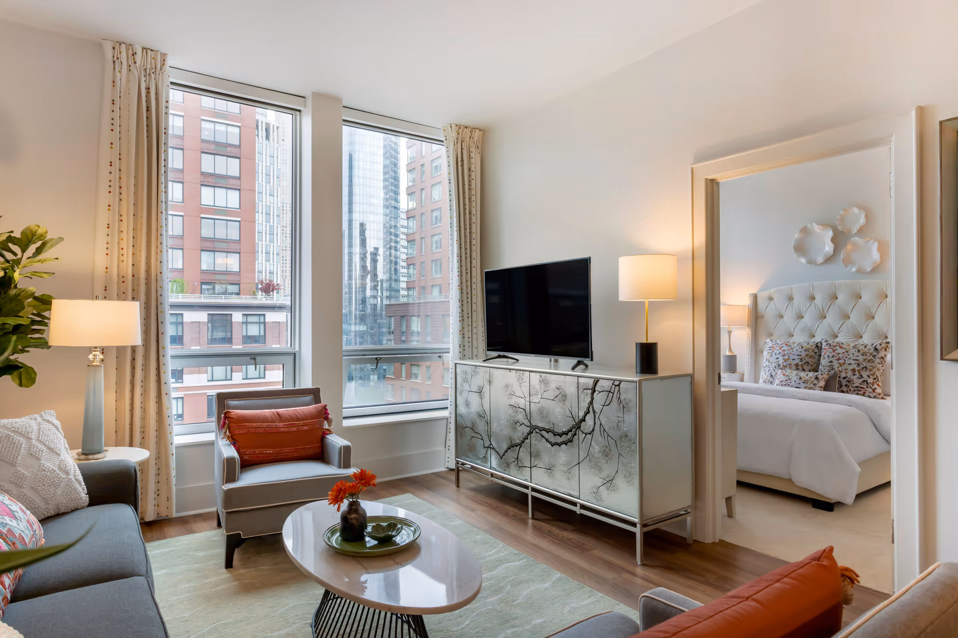A modern living room with large windows showing city buildings outside. The room features a gray sofa with colorful pillows, a gray armchair with an orange cushion, a round coffee table with a flower vase, a decorative cabinet with a flat-screen TV on top, and two lamps. Through an open doorway, a bedroom with a tufted headboard and floral pillows is visible.