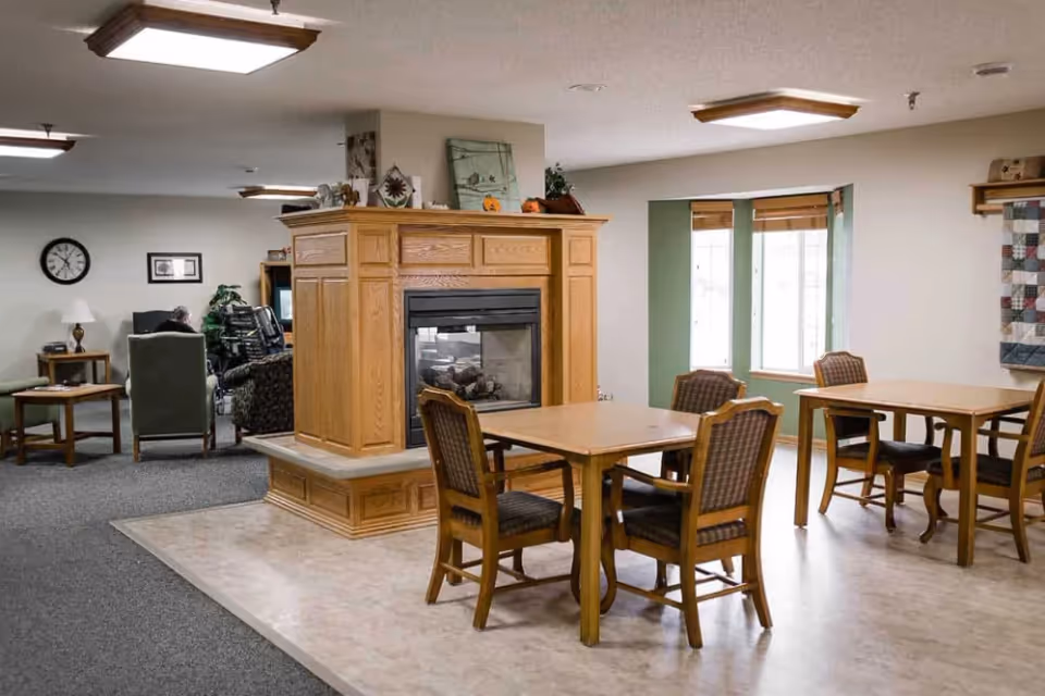 A senior living facility common area featuring a central wooden fireplace with decorative items on top, surrounded by tables and chairs. There are windows with natural light coming in, a clock on the wall, and seating areas with armchairs in the background.