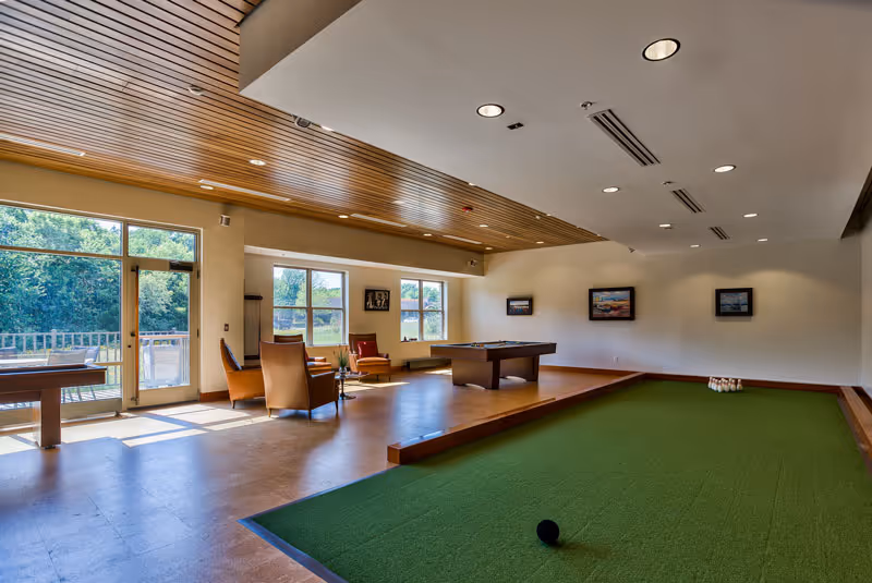 A spacious recreational room with a large indoor bocce ball court in the foreground, a pool table in the middle, and several chairs arranged near large windows that let in natural light. The ceiling has wooden paneling and recessed lighting, and there are framed pictures on the walls. A door leads to an outdoor area with greenery visible outside.
