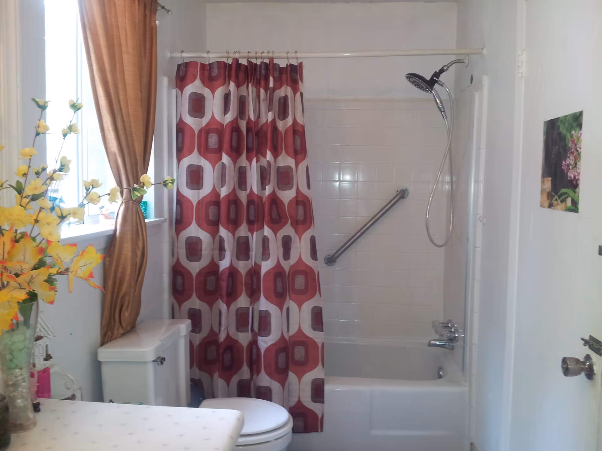 Bathroom with a white bathtub and shower combination featuring a red and white patterned shower curtain. There is a silver grab bar on the tiled wall inside the shower. A toilet is positioned next to the bathtub, and a window with brown curtains is above the toilet. A vase with yellow flowers is on the counter beside the toilet. A small picture is hung on the white wall near the door.