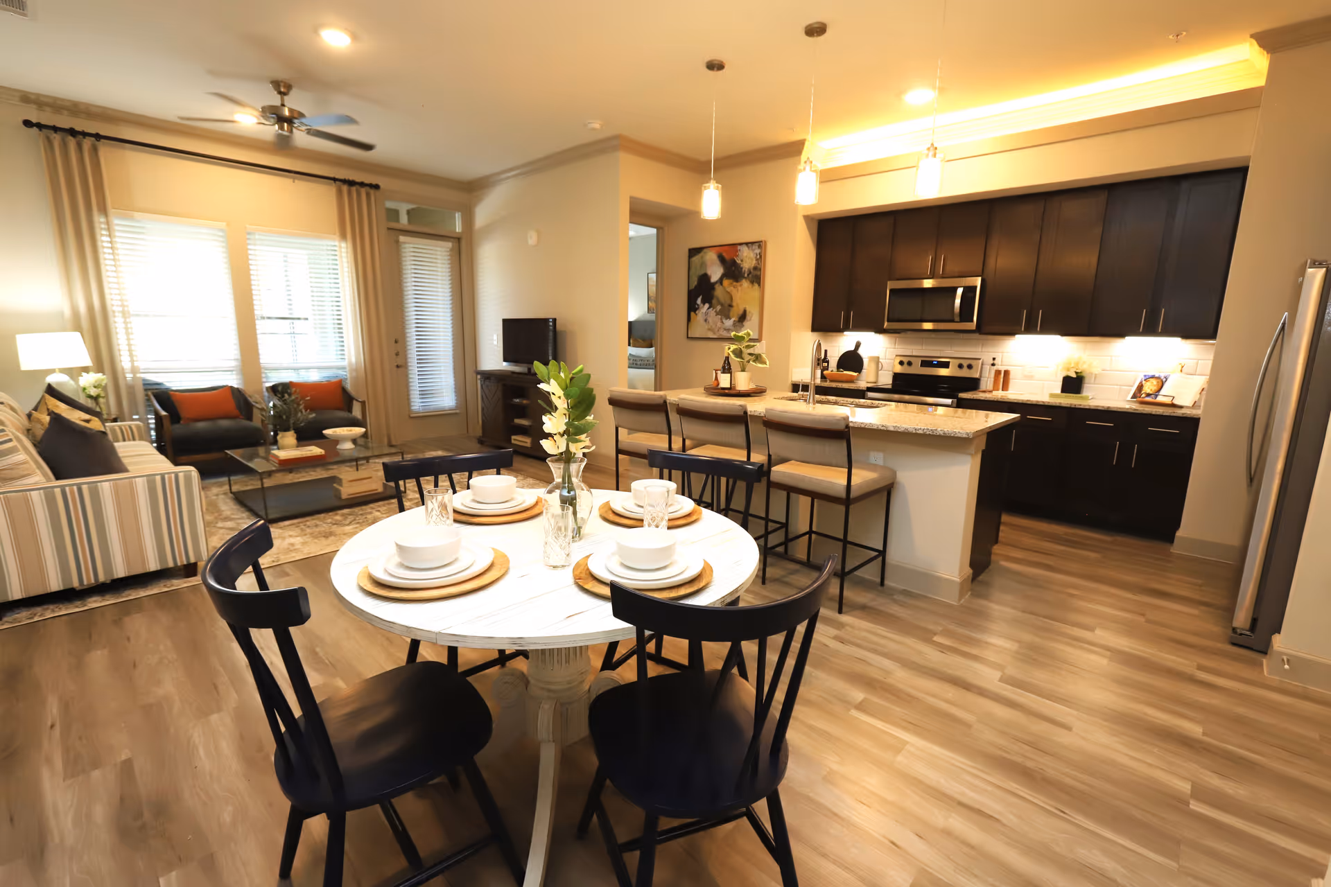 A bright and modern open-concept living space featuring a dining area with a round table set for four, a kitchen with dark wood cabinets, stainless steel appliances, and a breakfast bar with three stools. The living room area includes a striped sofa, two armchairs with orange cushions, a glass coffee table, and a TV on a wooden stand. Large windows with blinds and curtains allow natural light to fill the room.