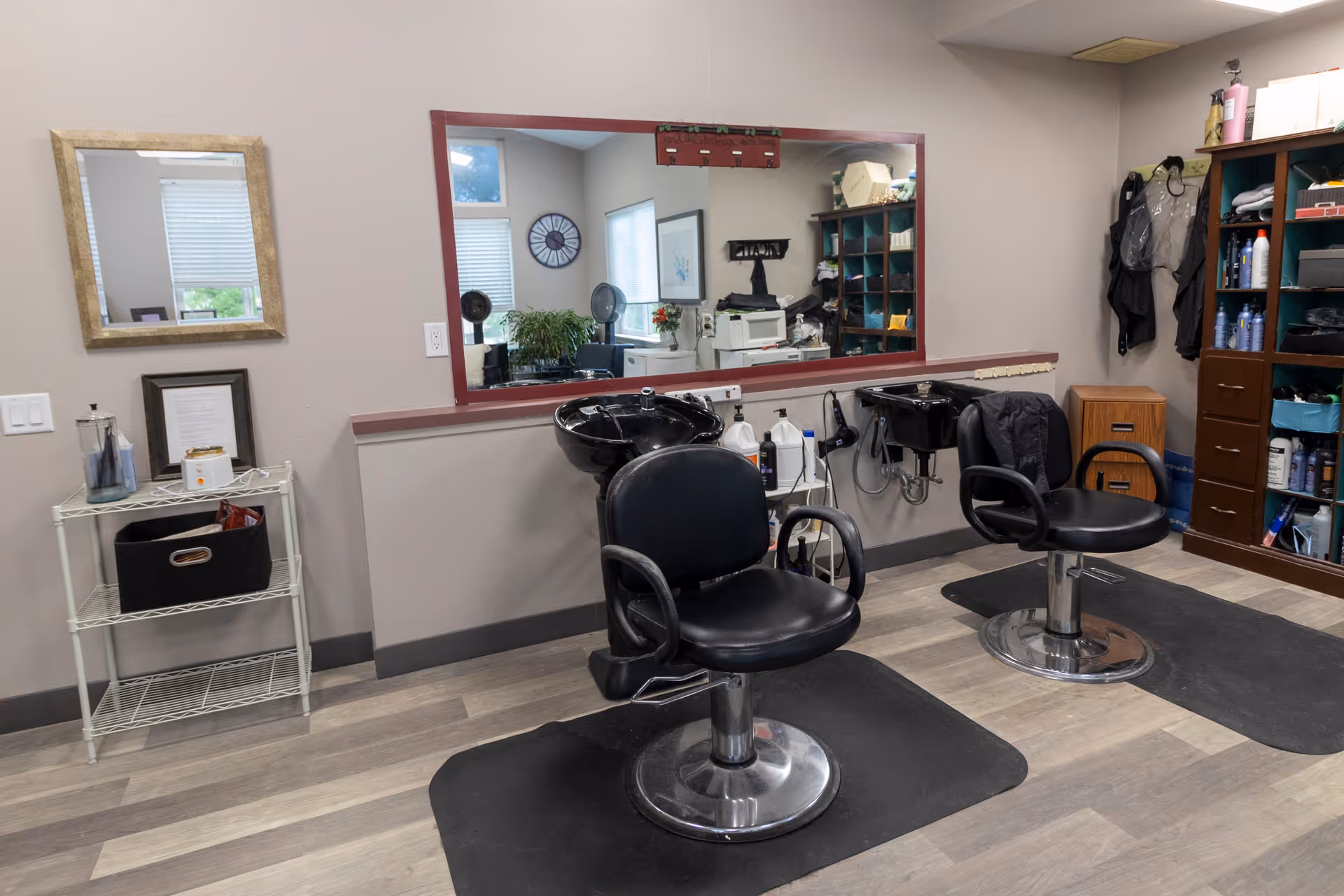 Interior view of a hair salon area with two black salon chairs in front of a large mirror. There are hair washing sinks behind the chairs, shelves with hair products and supplies, and a small table with a mirror and framed picture on the left side. The floor has a wood-like finish and there are black mats under the chairs.