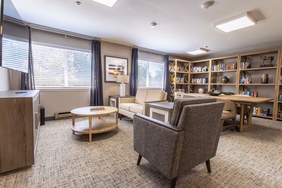 A well-lit living room area with a beige loveseat, a gray armchair, a round wooden coffee table, and a wooden side table with a lamp. Large windows with blinds and dark curtains allow natural light to fill the room. In the background, there is a large wooden bookshelf filled with books and decorative items, along with a wooden table and chairs for reading or activities.