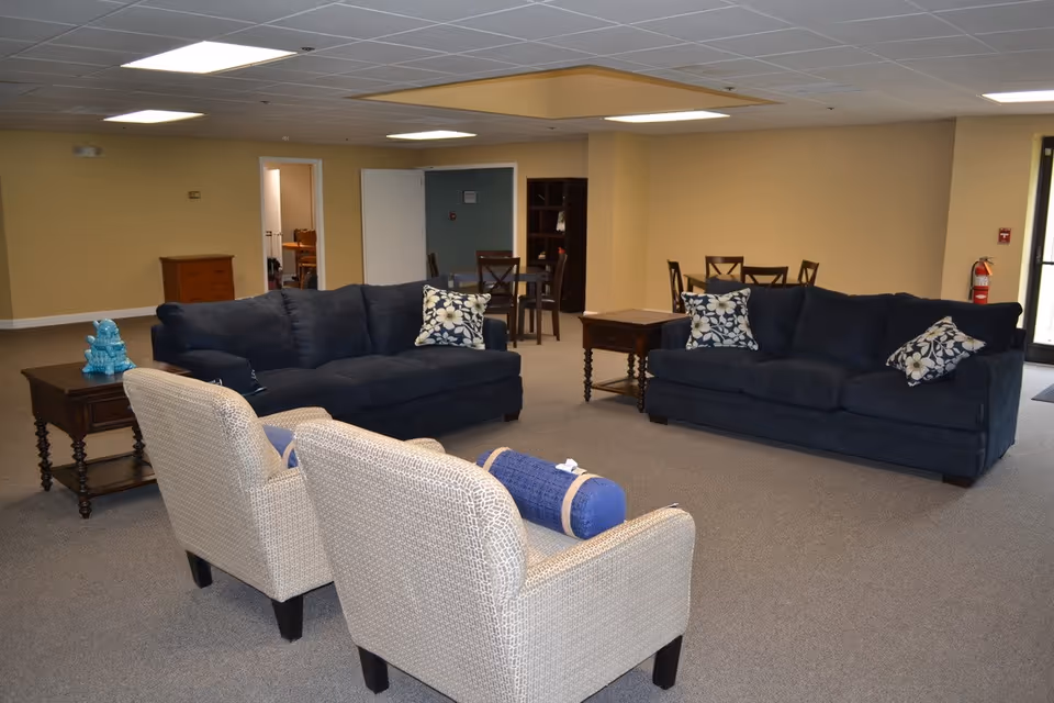A senior living facility common area with two dark blue sofas adorned with floral pillows, two beige armchairs with a blue bolster pillow, wooden side tables, and a dining table with chairs in the background. The room has beige walls, a gray carpet, and a drop ceiling with fluorescent lights.