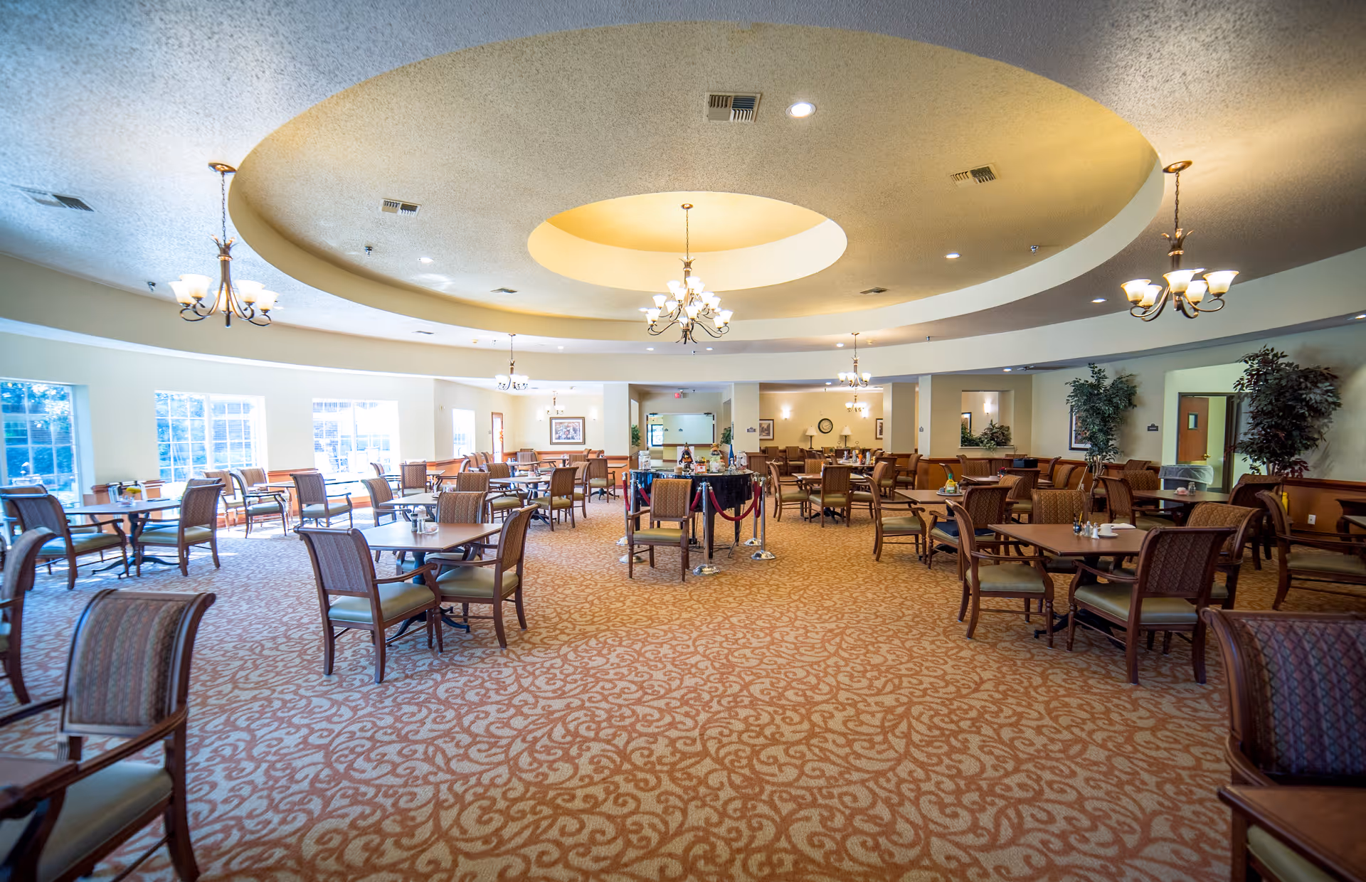 A spacious dining room with multiple wooden tables and chairs arranged neatly on a patterned carpet. The ceiling features a large circular recessed design with chandeliers hanging down, providing warm lighting. Large windows on the left side allow natural light to fill the room. The room is decorated with plants and framed artwork on the walls.