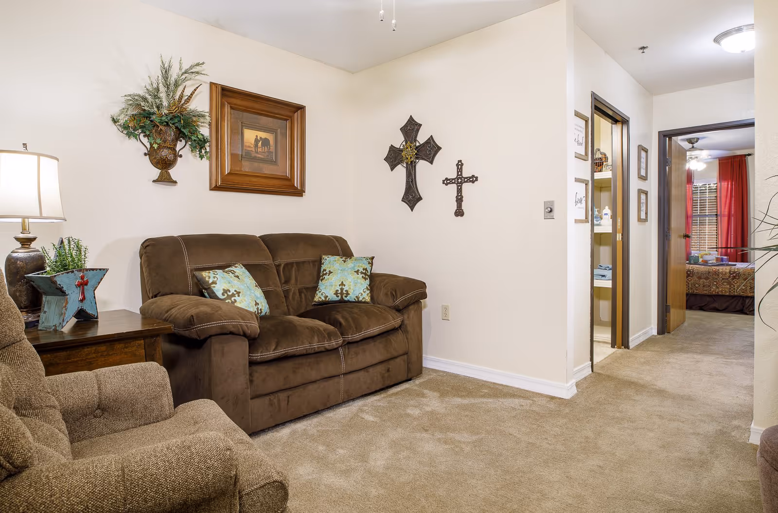 A cozy living room area with a brown loveseat and a matching armchair. The loveseat has two decorative pillows with a blue and brown pattern. On the wall above the loveseat are two decorative crosses and a framed picture. A side table with a lamp and a decorative star planter is next to the loveseat. The room has beige carpet and cream-colored walls. In the background, there is a hallway leading to a bedroom with red curtains and a ceiling fan.